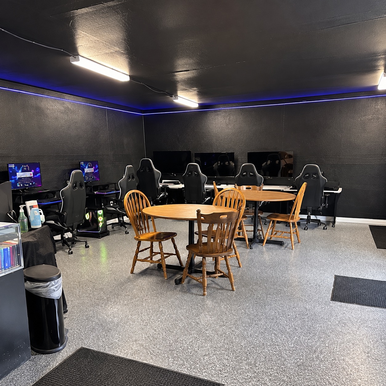 Commercial gaming room with a gray flake-coated floor beneath gaming PCs, racing-style chairs and round wooden tables, black walls with blue LED trim