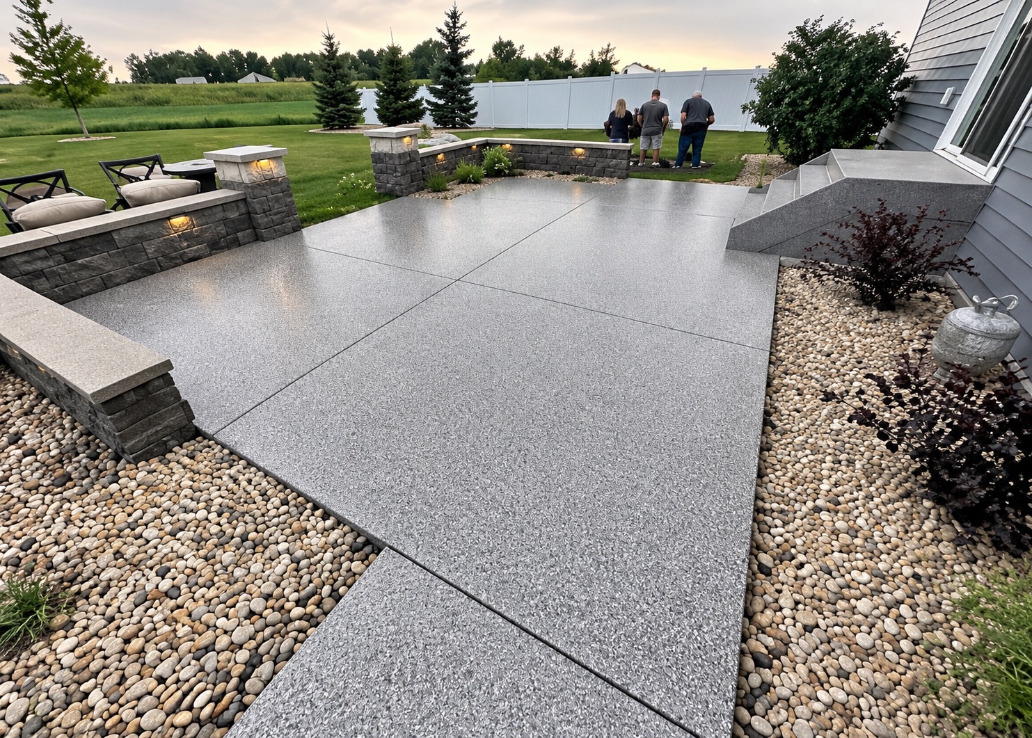 Backyard patio with a gray flake coating across the slab, walkway and entry steps, framed by lit stone retaining walls and river rock
